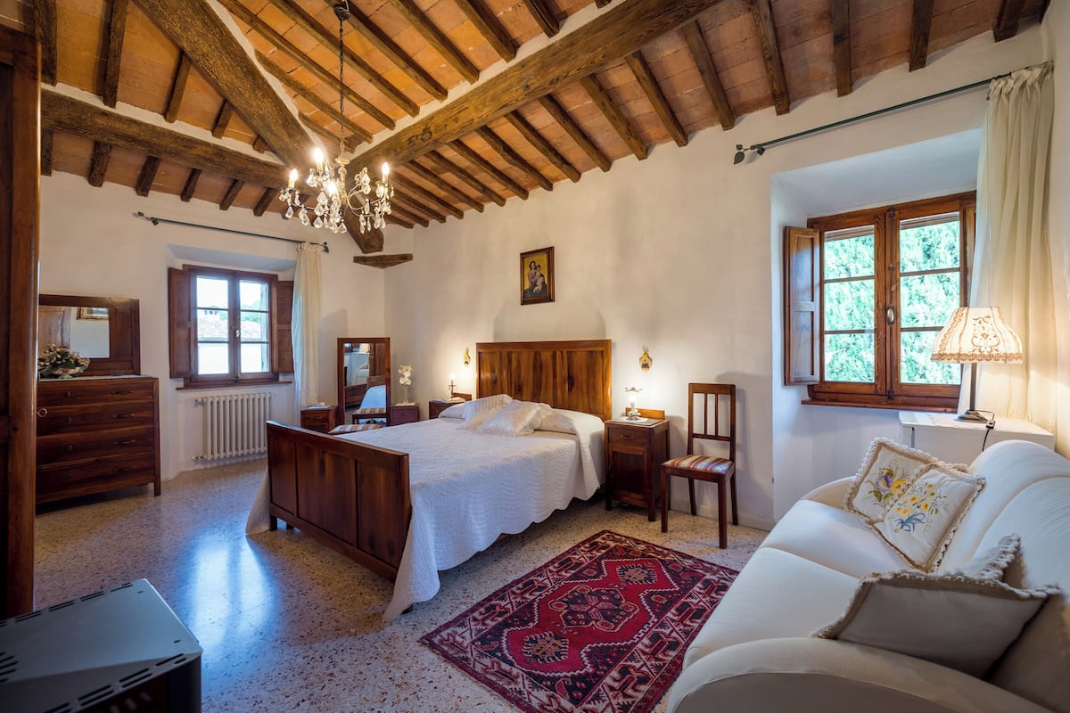 Une chambre avec un plafond en bois, un lit, une table de chevet, une commode, une chaise et une fenêtre avec des rideaux.