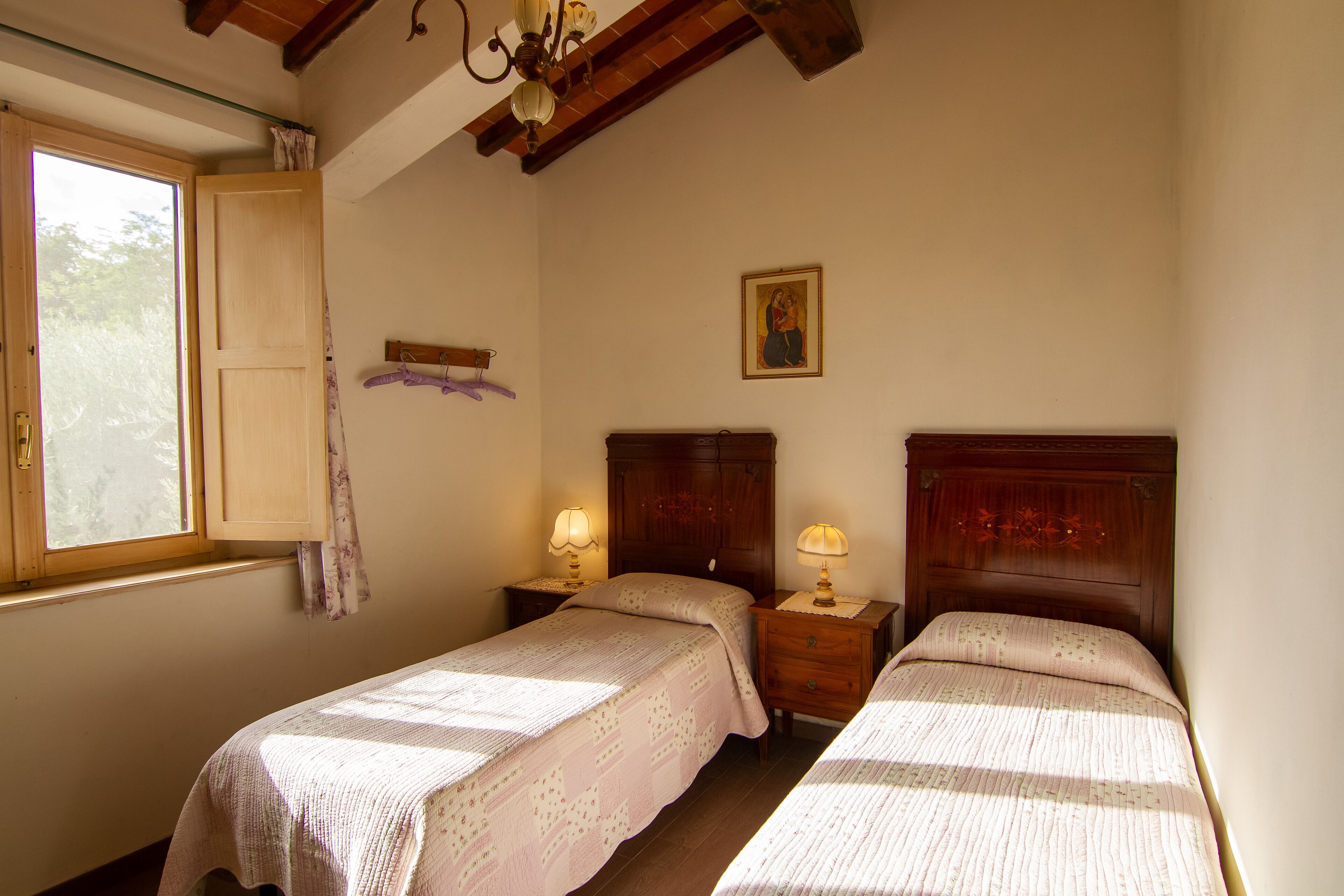 A bedroom with two single beds, wooden headboards, a nightstand with a lamp, a window with curtains, and a wall-mounted picture.
