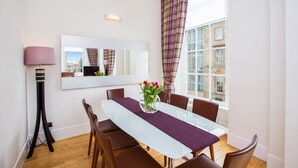 A dining area with a glass table, purple runner, and brown chairs. There is a vase with flowers on the table. A lamp and a mirror are on the wall. - Blythswood Square Apartments (Glasgow)