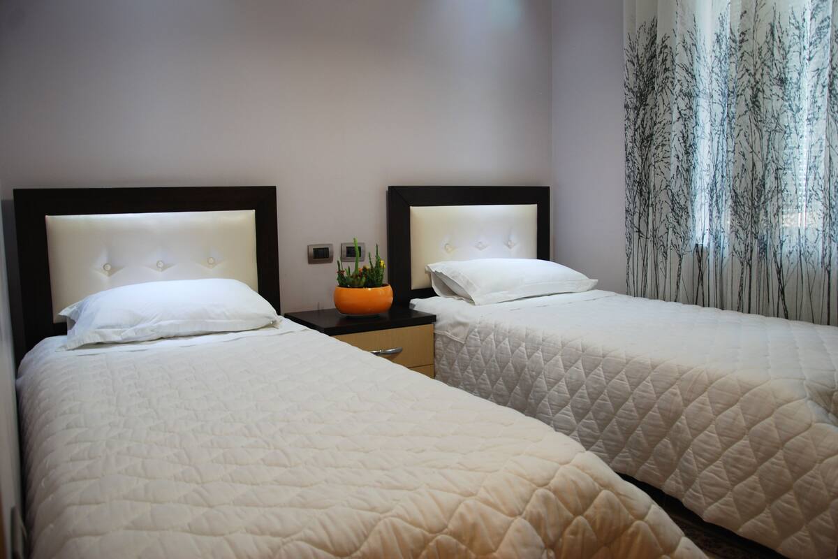A hotel room with two beds, a nightstand with a plant, and a window with sheer curtains.