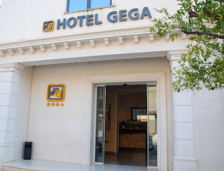 The exterior of Hotel Gega with a clear entrance, a logo with stars, and a visible clock on the wall.