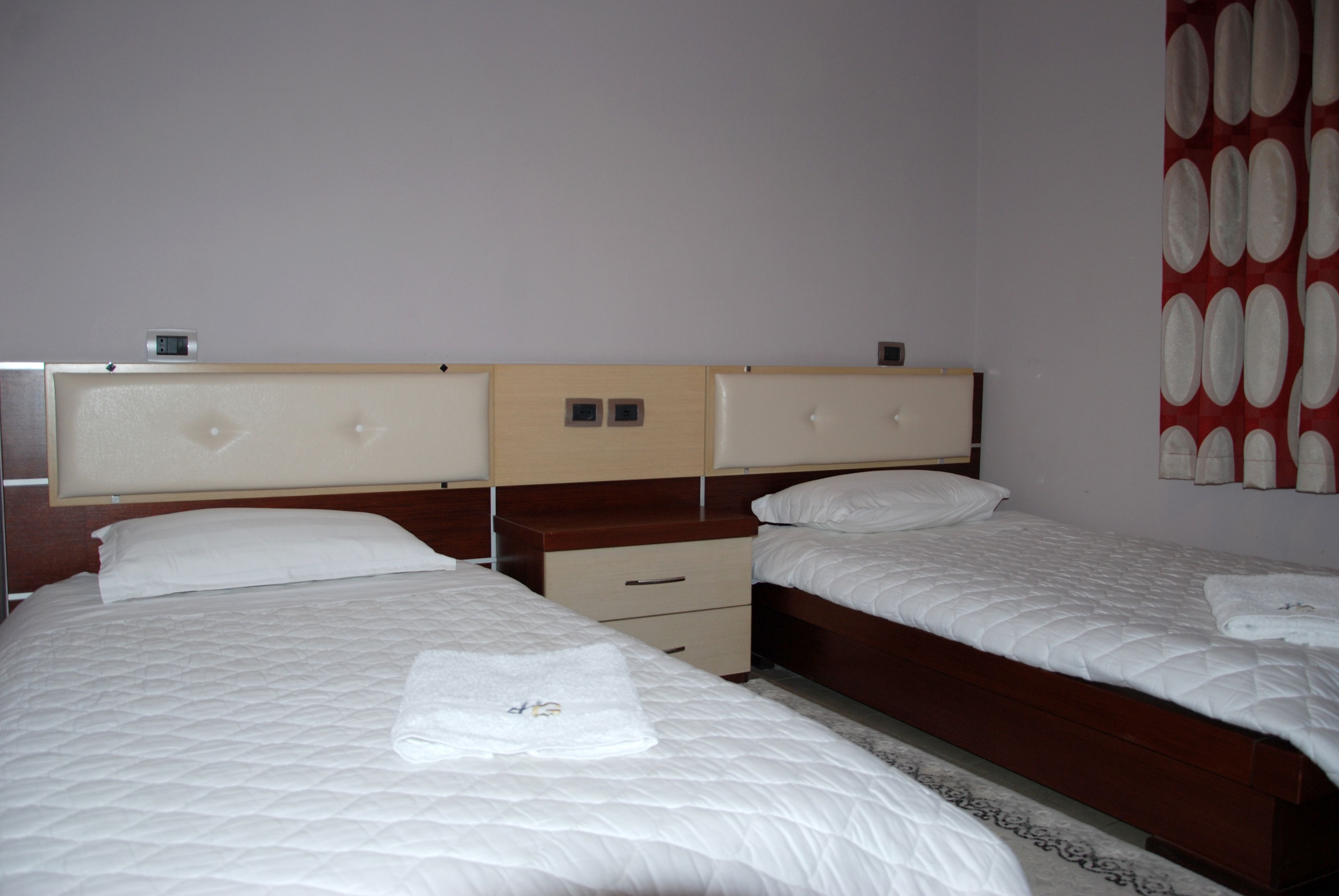 Quarto de hotel com duas camas, uma cabeceira de madeira, uma pequena mesa de cabeceira e uma cortina vermelha estampada.