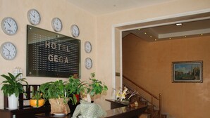 A hotel reception area with multiple clocks displaying different city times, a large digital clock, and a staircase leading to another floor. - Hotel Gega (Berat)