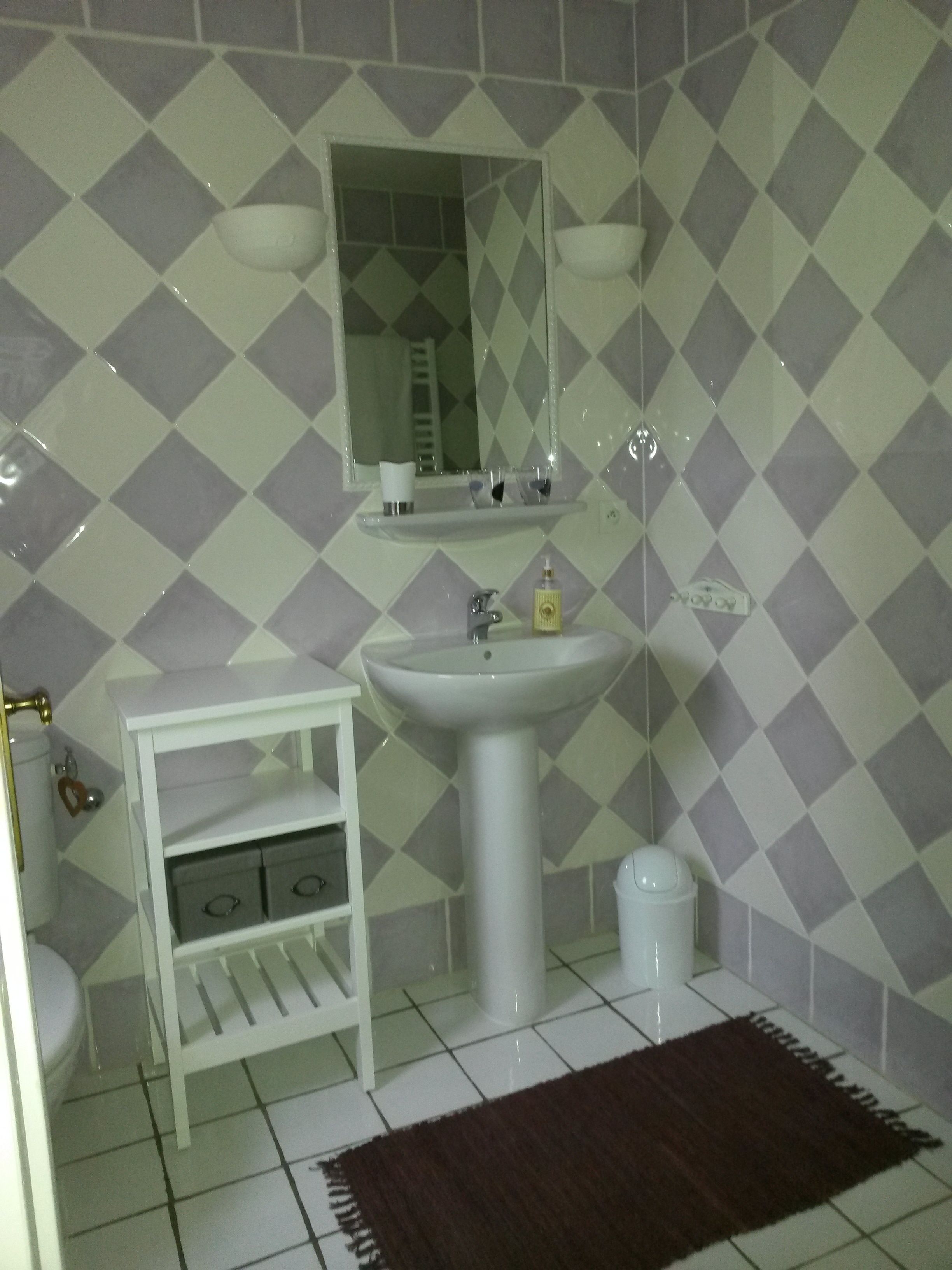 Une salle de bain avec un lavabo blanc, un miroir et des murs carrelés. On y trouve une étagère blanche avec des tiroirs et une petite poubelle.