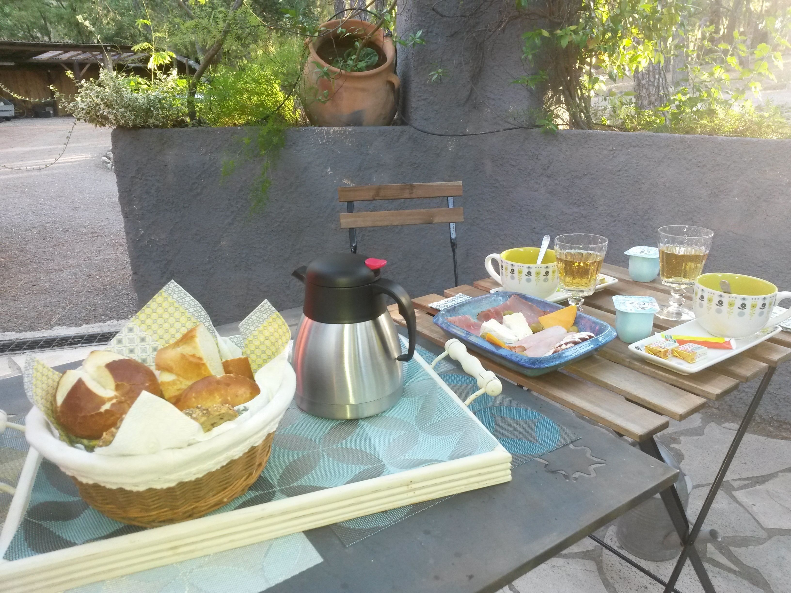 Desayuno con pan, queso, jamón y una cafetera sobre una mesa al aire libre.
