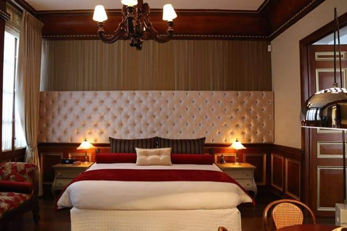Habitación de hotel con una cama grande, dos mesitas de noche con lámparas, un sillón tapizado de color rojo y una araña.
