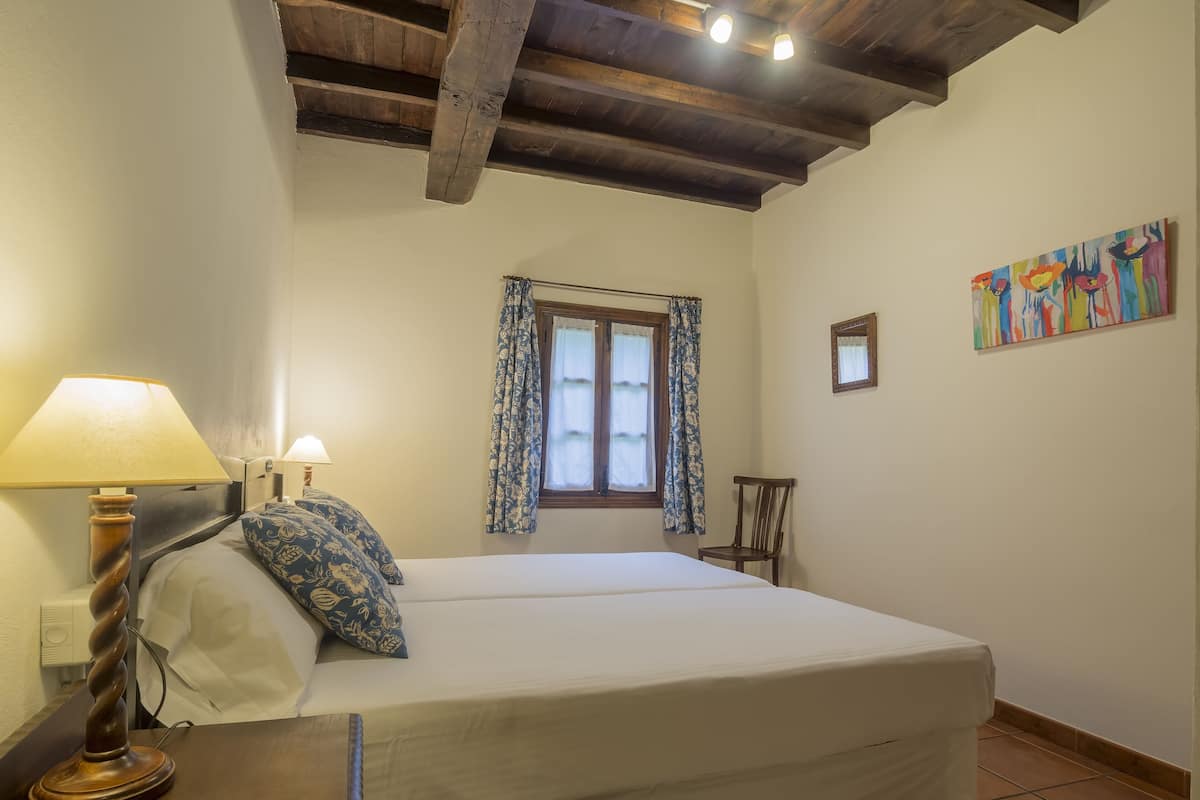 Quarto com cama, abajur, cadeira, janela com cortinas e um quadro na parede.