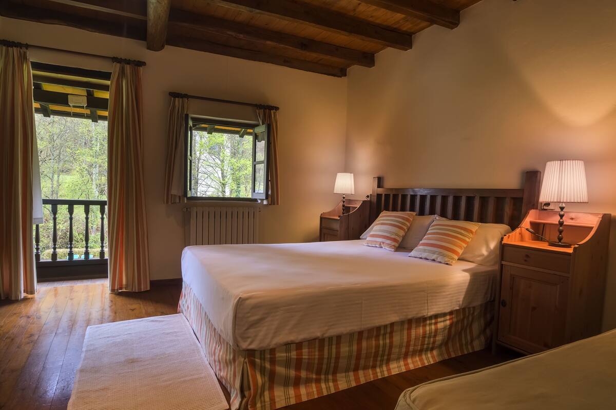 Quarto com cama de madeira, duas mesas de cabeceira com abajures, janela com cortinas e vista para o verde lá fora.