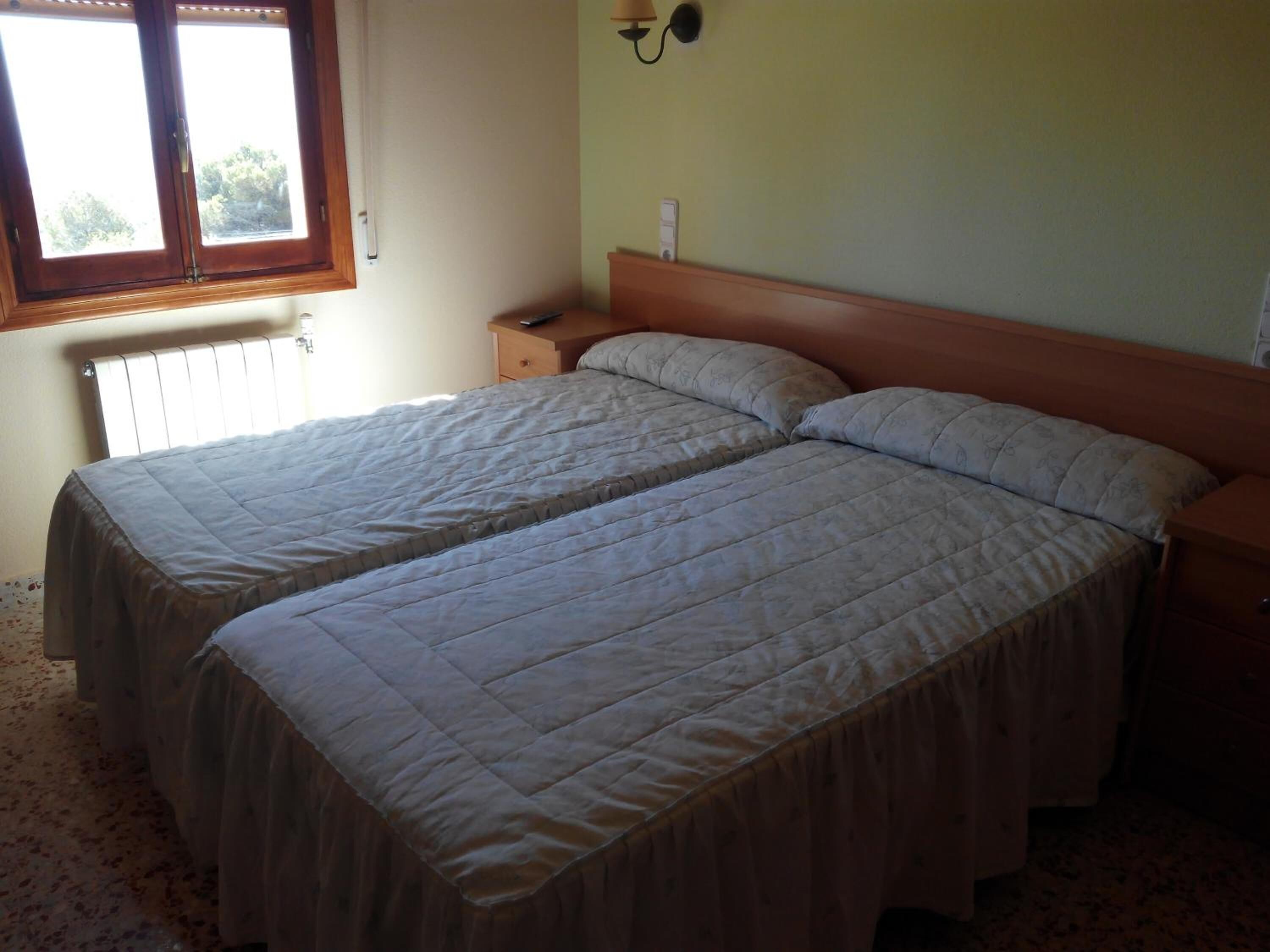 Une chambre avec deux lits simples, une table de chevet, une lampe fixée au mur et une fenêtre avec des stores.