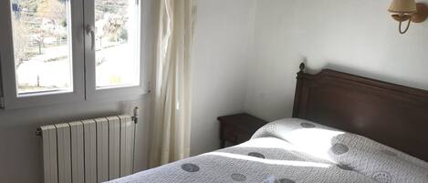 A bedroom with a bed, a nightstand, a window with curtains, a radiator, and a wall-mounted lamp.