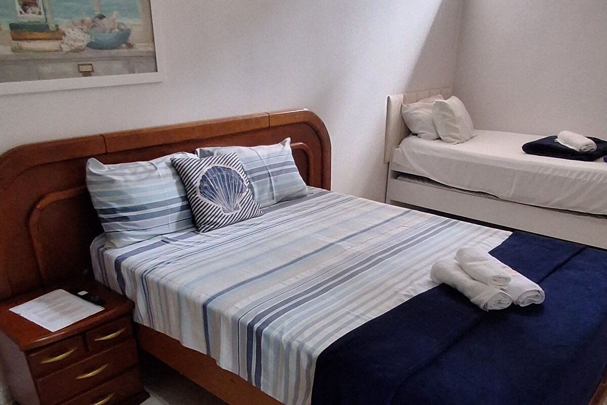 Un dormitorio con cama, mesita de noche, ventana y un cuadro en la pared.