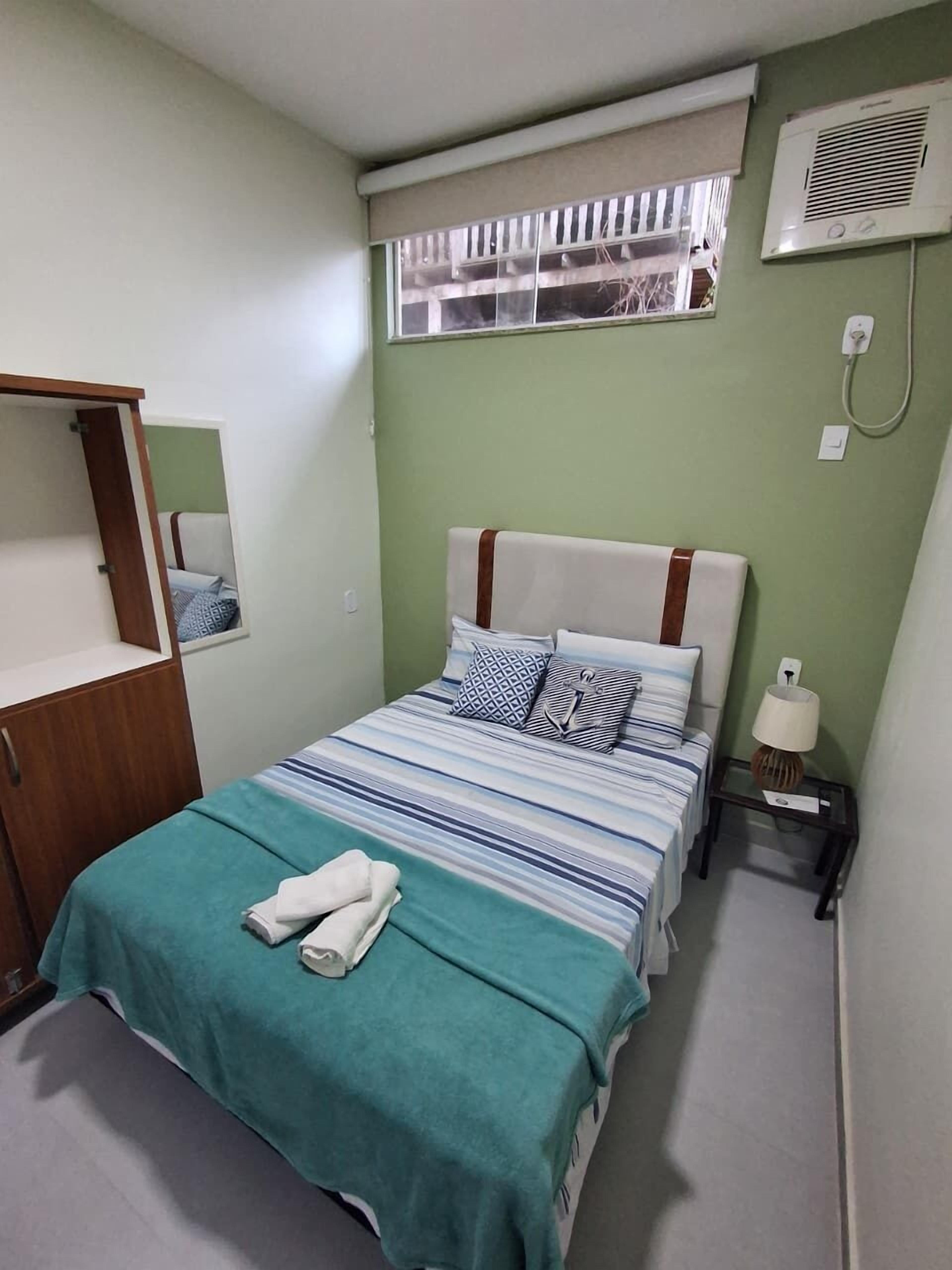A bedroom with a bed, a wardrobe, a window with blinds, an air conditioning unit, and a bedside lamp.