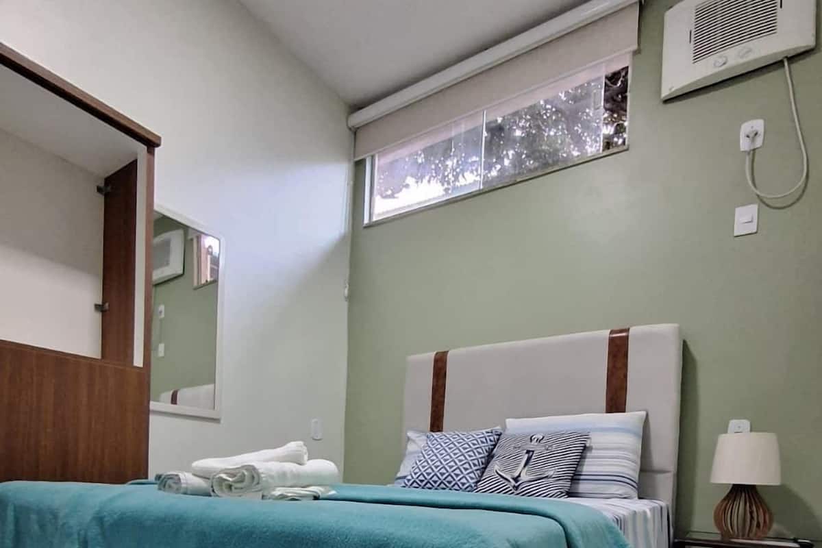 Un dormitorio con cama, mesita de noche, lámpara, ventana y espejo.