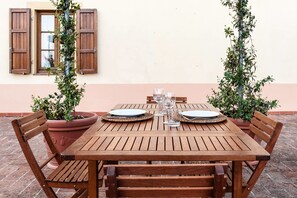 A wooden table set for two with plates, cutlery, and glasses, surrounded by wooden chairs, with potted plants and a building in the background. - Tenuta degli Obizzi (Montecarlo)