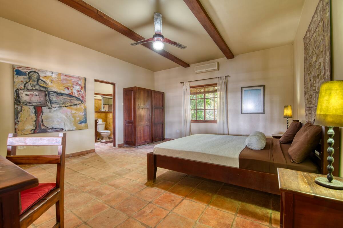 Ein Schlafzimmer mit einem Bett, Holzmöbeln, einem Deckenventilator, einem Fenster mit Vorhängen und einem Bild an der Wand.