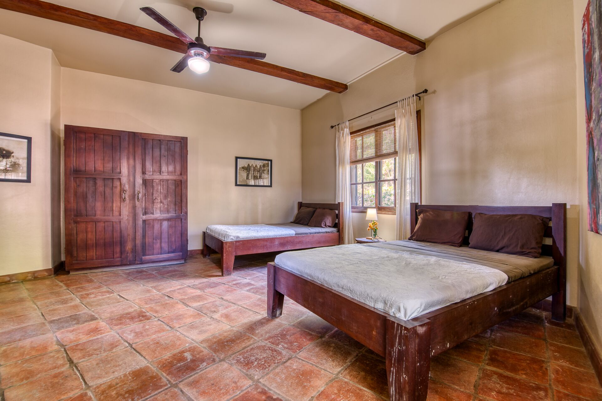 Une chambre avec un grand lit, un plus petit lit, une armoire en bois, un ventilateur de plafond et une fenêtre avec des rideaux.