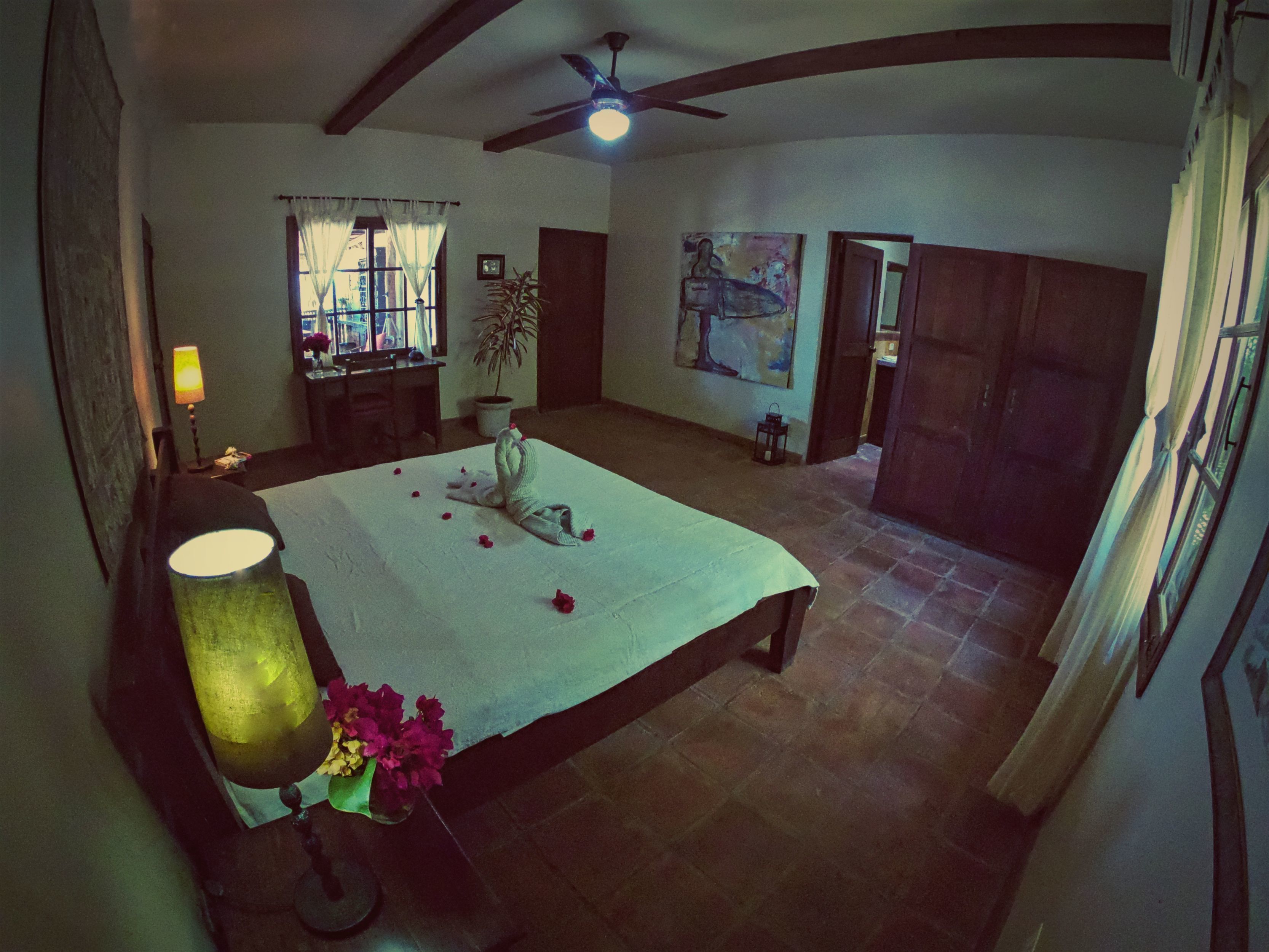 A spacious bedroom with a large bed, a ceiling fan, a window with curtains, a potted plant, and a painting on the wall.