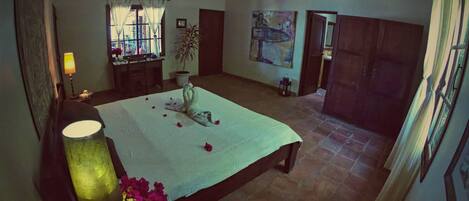 A spacious bedroom with a large bed, a ceiling fan, a window with curtains, a potted plant, and a painting on the wall.