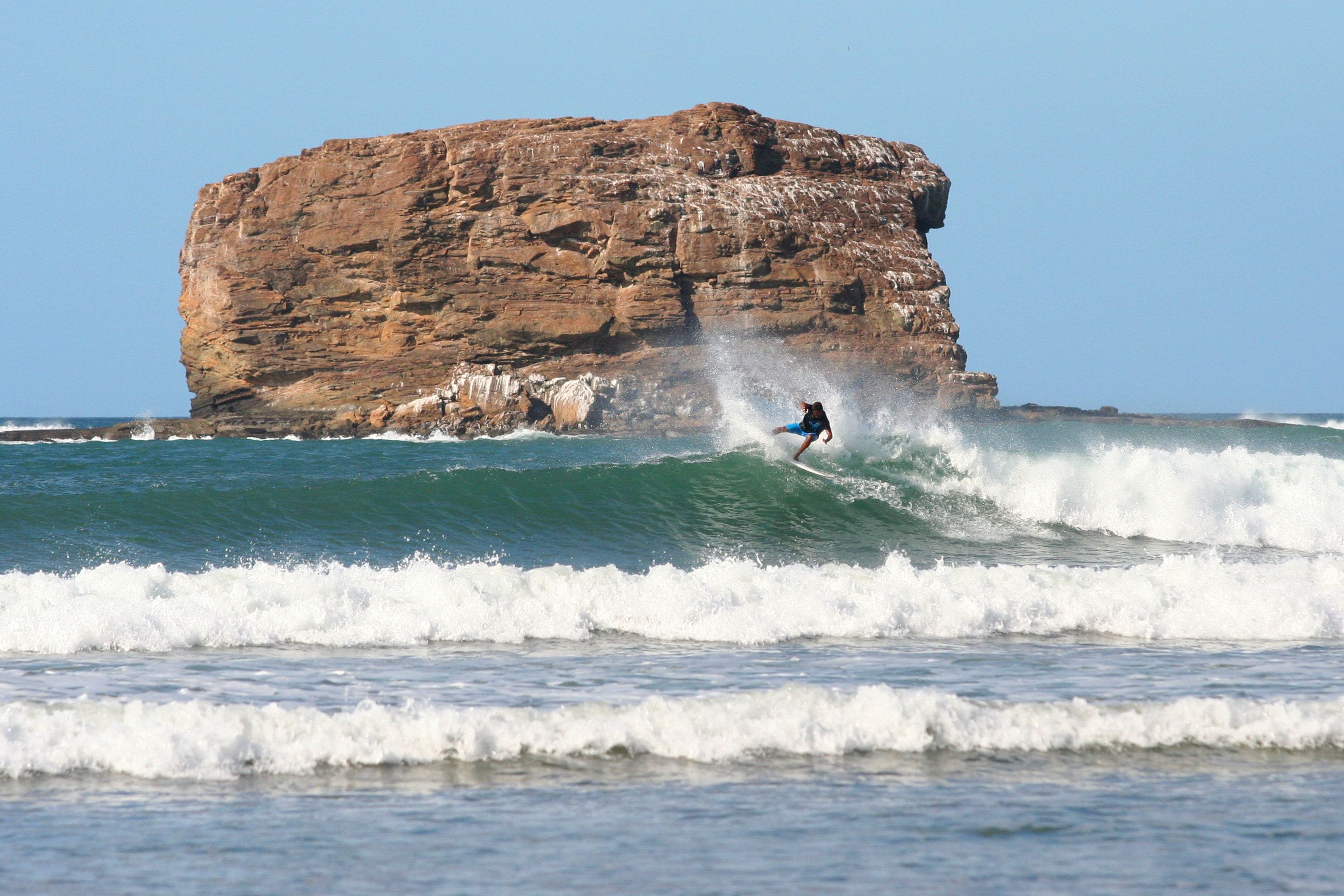 A surfer riding a wave near a large rock formation.