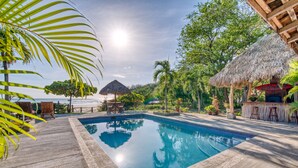 Une piscine dotée d’une terrasse en bois, entourée de plantes tropicales et bordée d’une structure à toit de chaume.