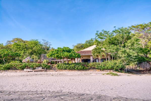 A beachfront property with a thatched-roof building, surrounded by lush greenery and a sandy beach.