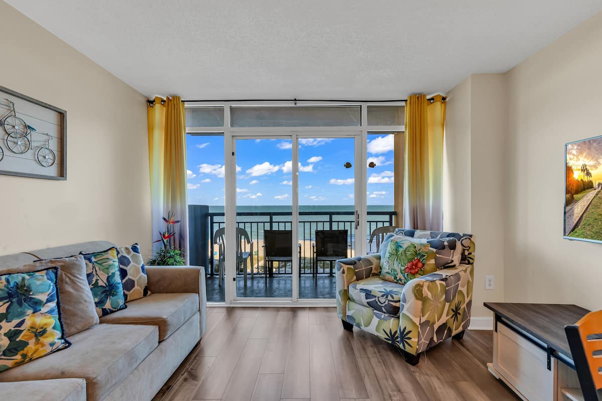 A living room with a sofa, armchair, and a dining table. There is a large window with curtains, and a view of the beach and sky.