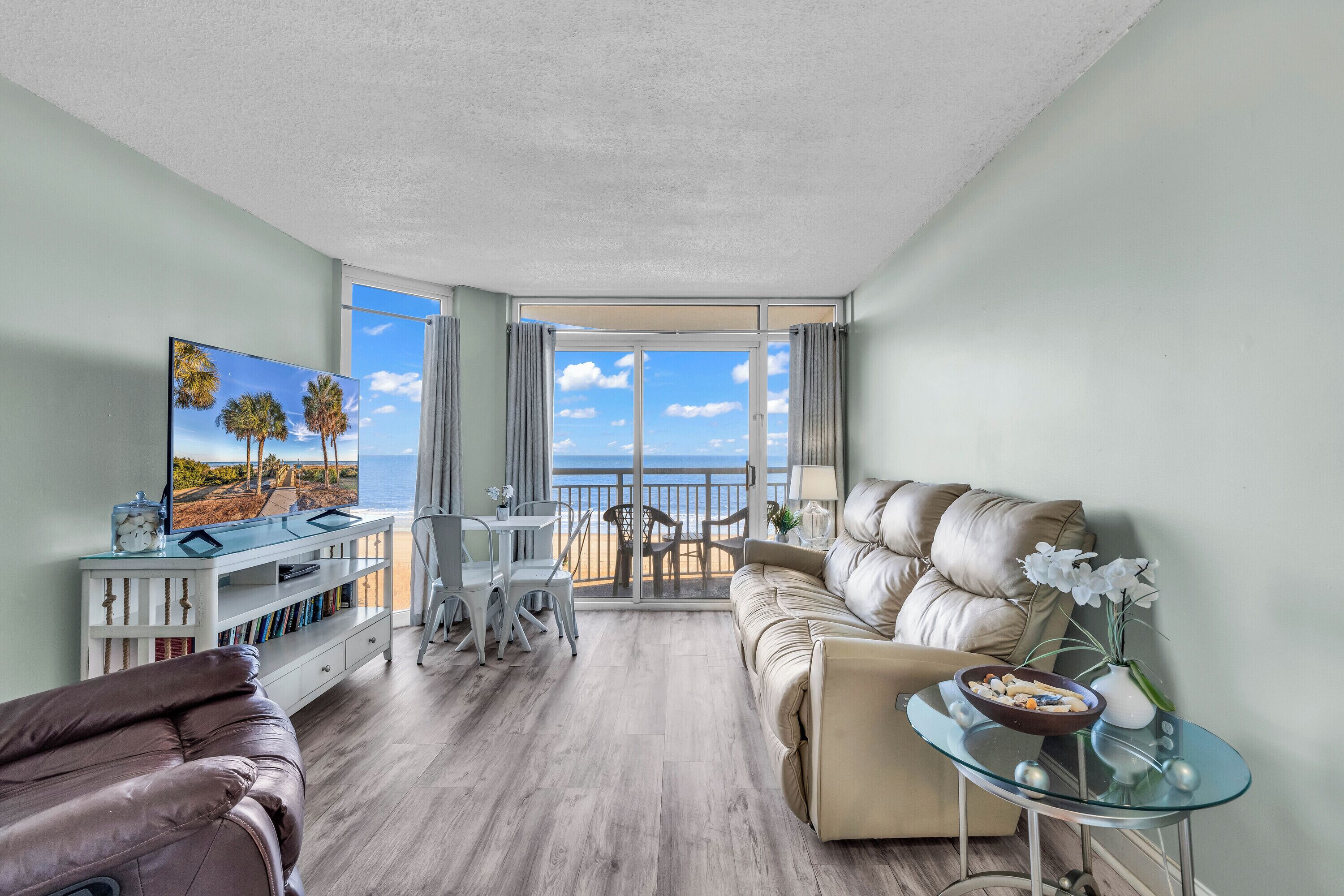A modern living room with a sofa, a glass coffee table, and a TV on a white console. A balcony with a view of the ocean is visible through the glass doors.