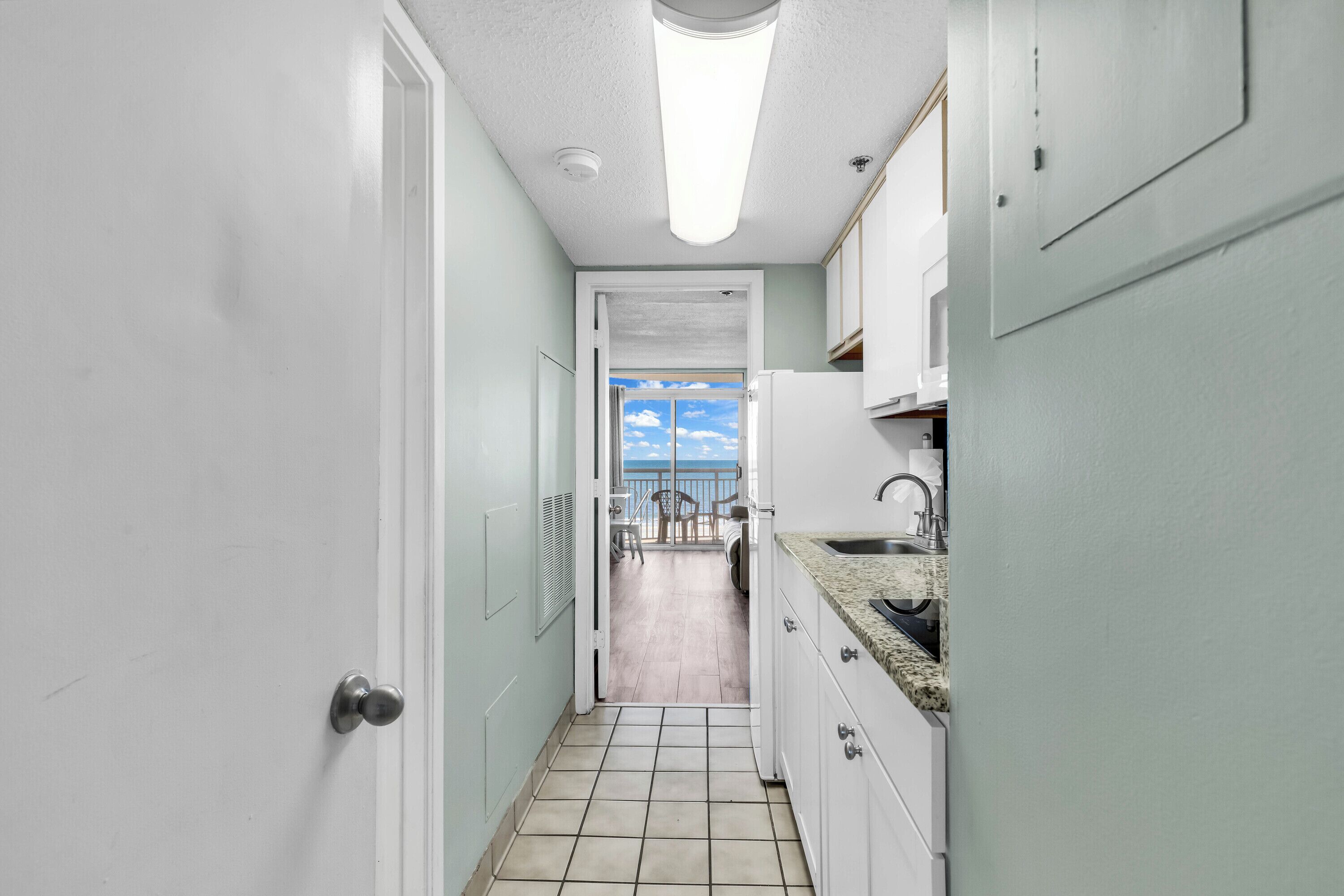 A narrow kitchen with white cabinets, a tiled floor, and a view to a balcony with a chair and table.