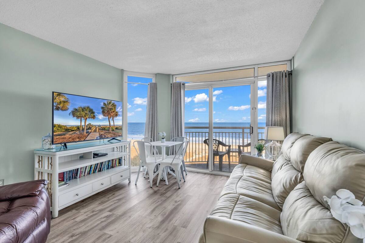 A living room with a TV, white furniture, a beige sofa, and a balcony with a view of the ocean.