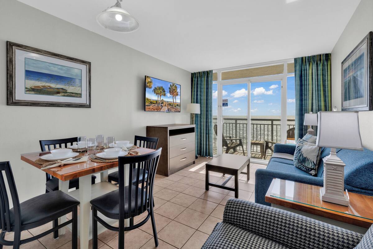 A dining area with a wooden table set for four, a sofa, and a balcony with a view.
