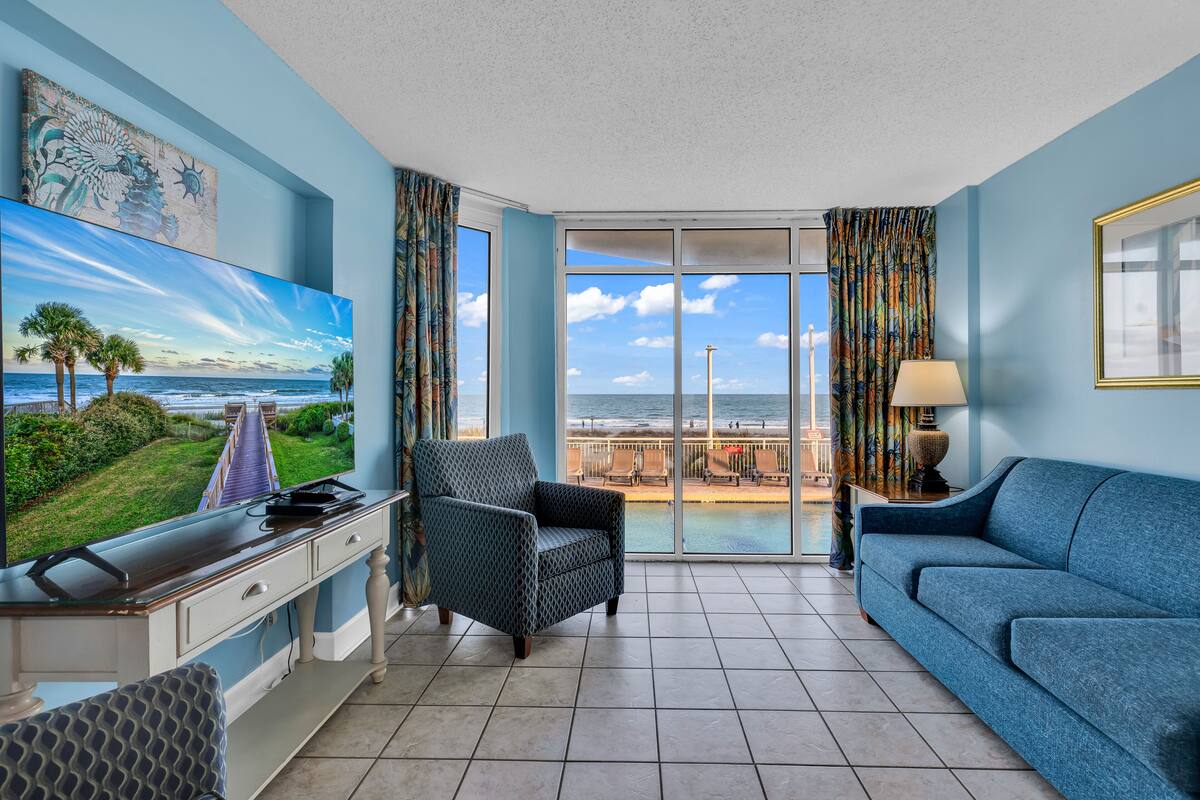 A living room with a large TV, a sofa, a chair, and a view of the ocean through a sliding glass door.
