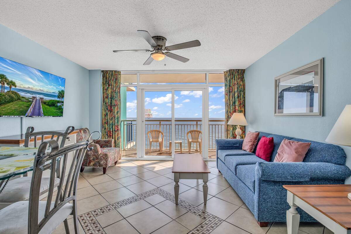 A living room with a blue sofa, a dining table, and chairs. There is a ceiling fan, a large window with a view of the sky, and a painting on the wall.