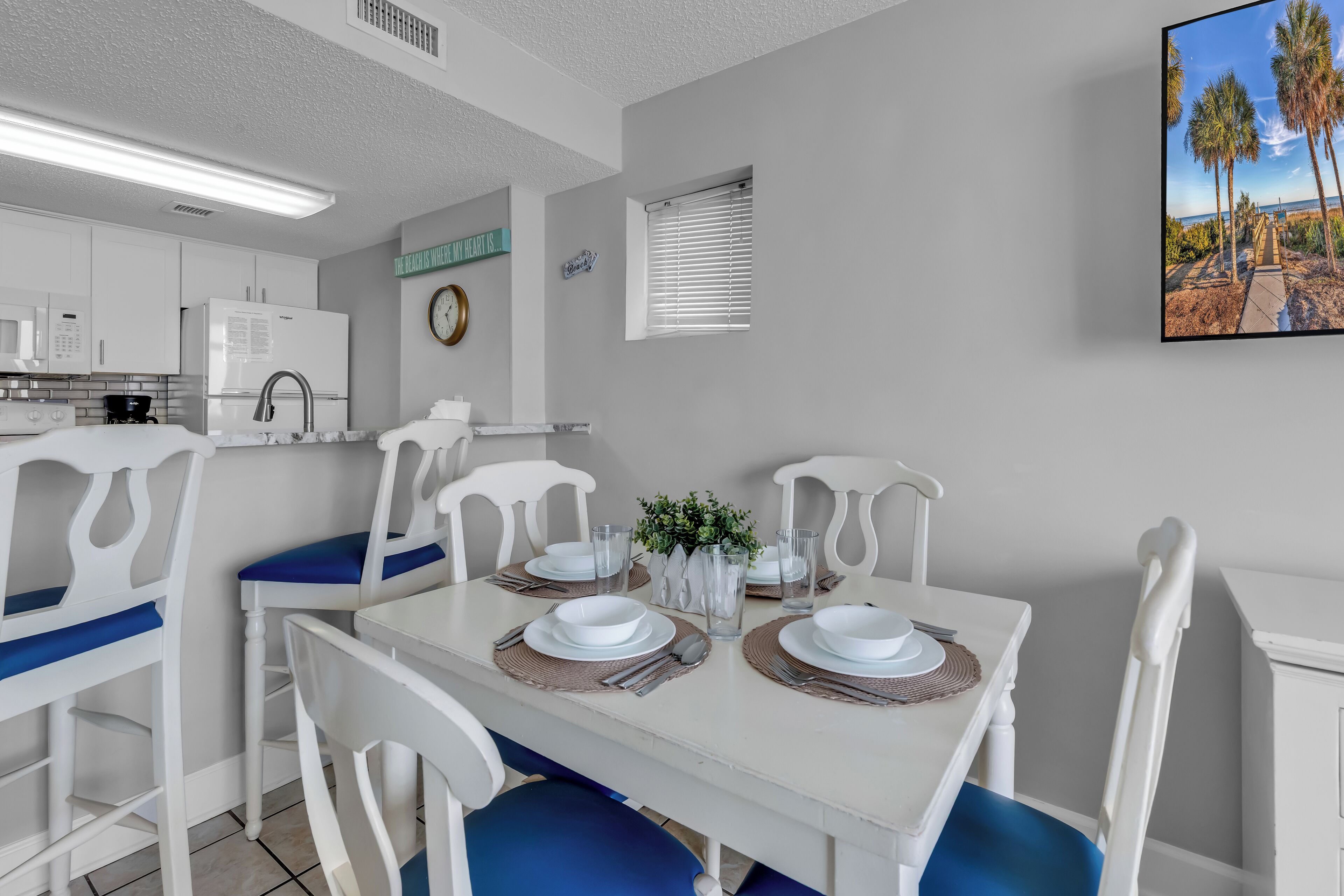 A dining area with a white table set for four, surrounded by white chairs with blue cushions. A small plant is on the table, and there is a clock on the wall.