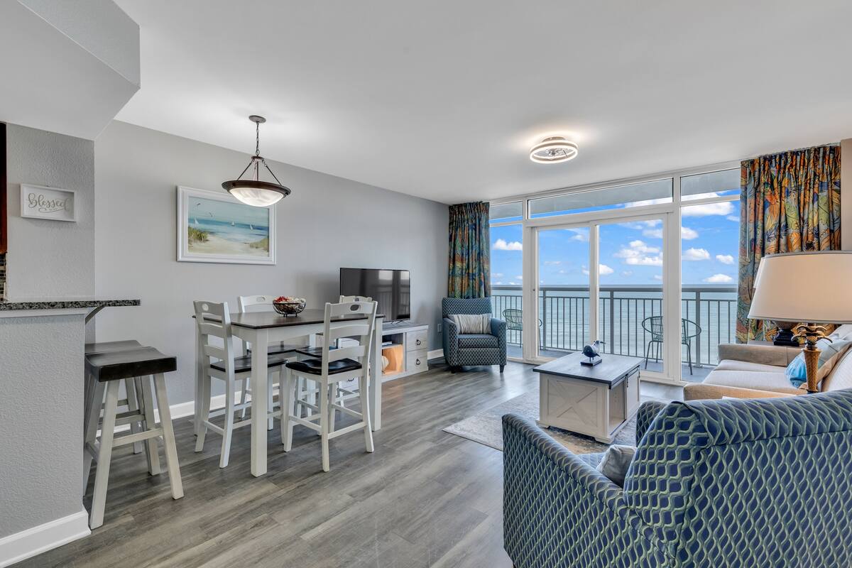 A modern living room with a dining area, a balcony with a view, and a kitchen area.