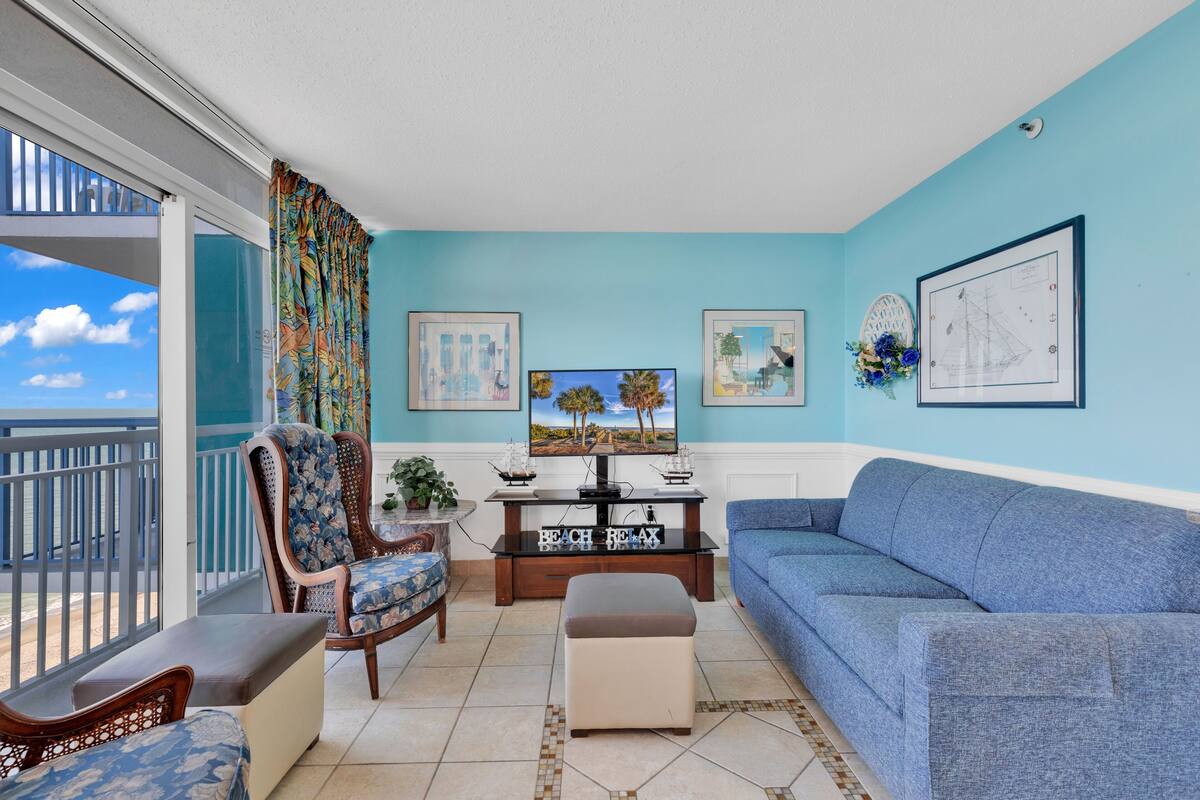 A living room with a blue sofa, a wooden chair, a coffee table, and a TV. There are framed pictures on the walls and a balcony with a railing.