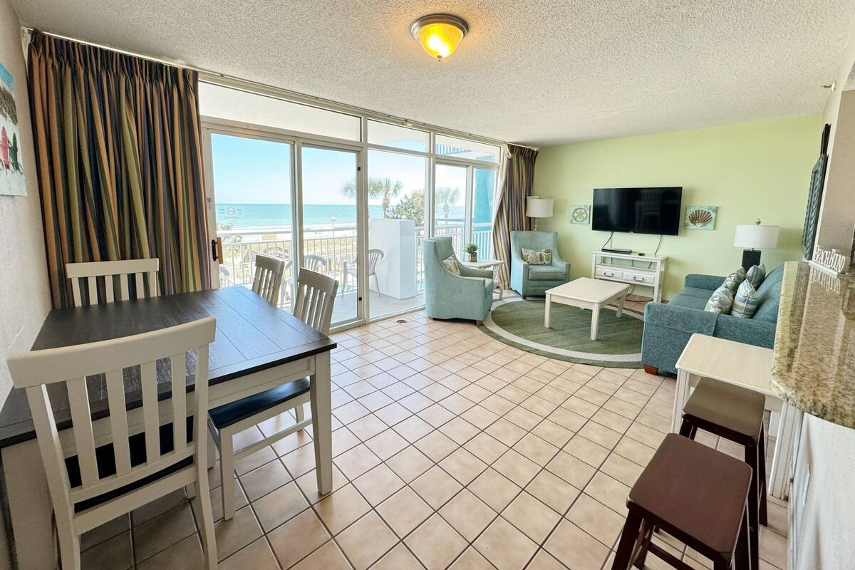 A living room with a dining area, a TV, and a balcony with a view of the sea.