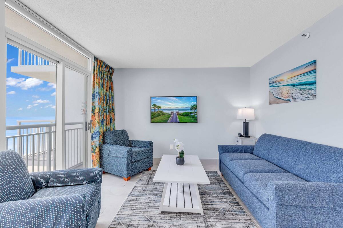 A modern living room with a balcony, a flat-screen TV, and a painting of a beach scene.