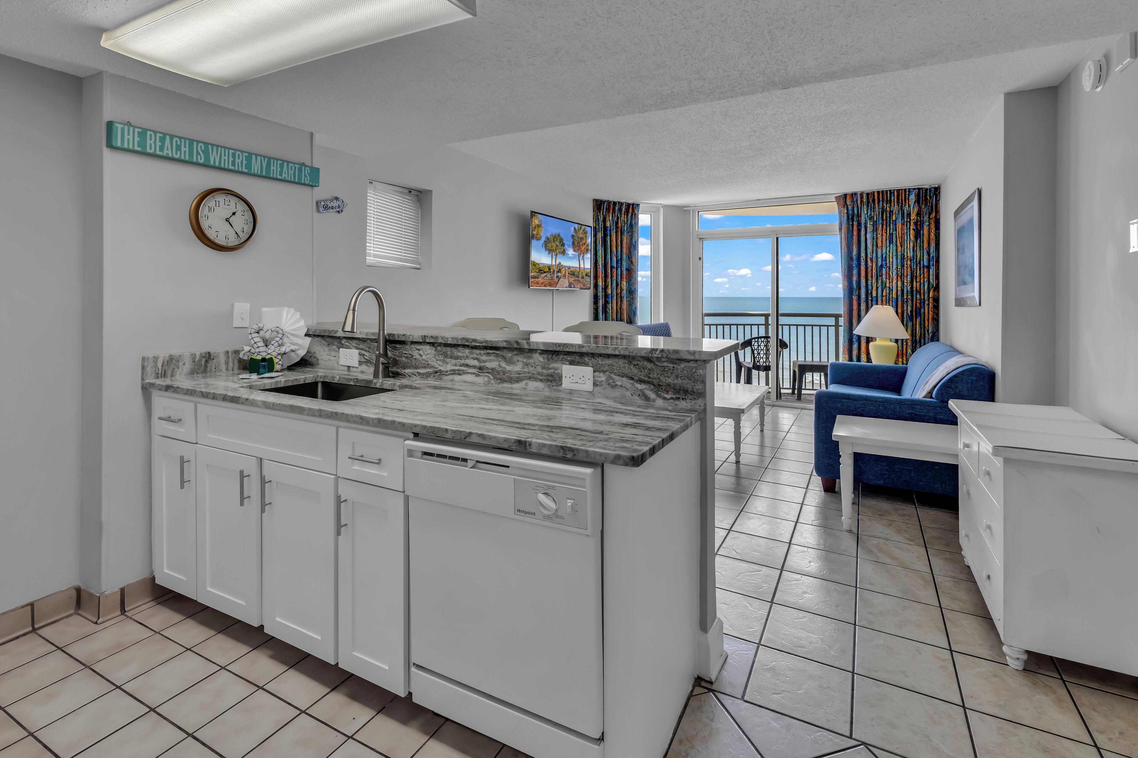A modern kitchen with white cabinets, a built-in dishwasher, and a marble countertop. There is a dining area with a blue couch and a view of the ocean.