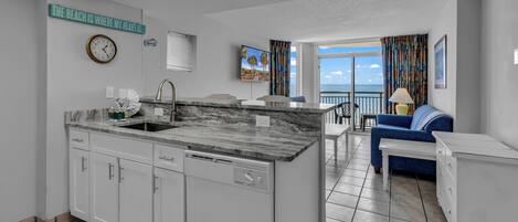 A modern kitchen with white cabinets, a built-in dishwasher, and a marble countertop. There is a dining area with a blue couch and a view of the ocean.