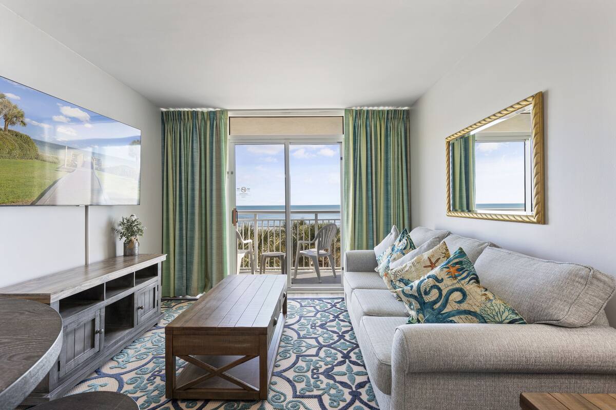 A modern living room with a sofa, a coffee table, and a TV on a wall. There is a balcony with a view of the sea.