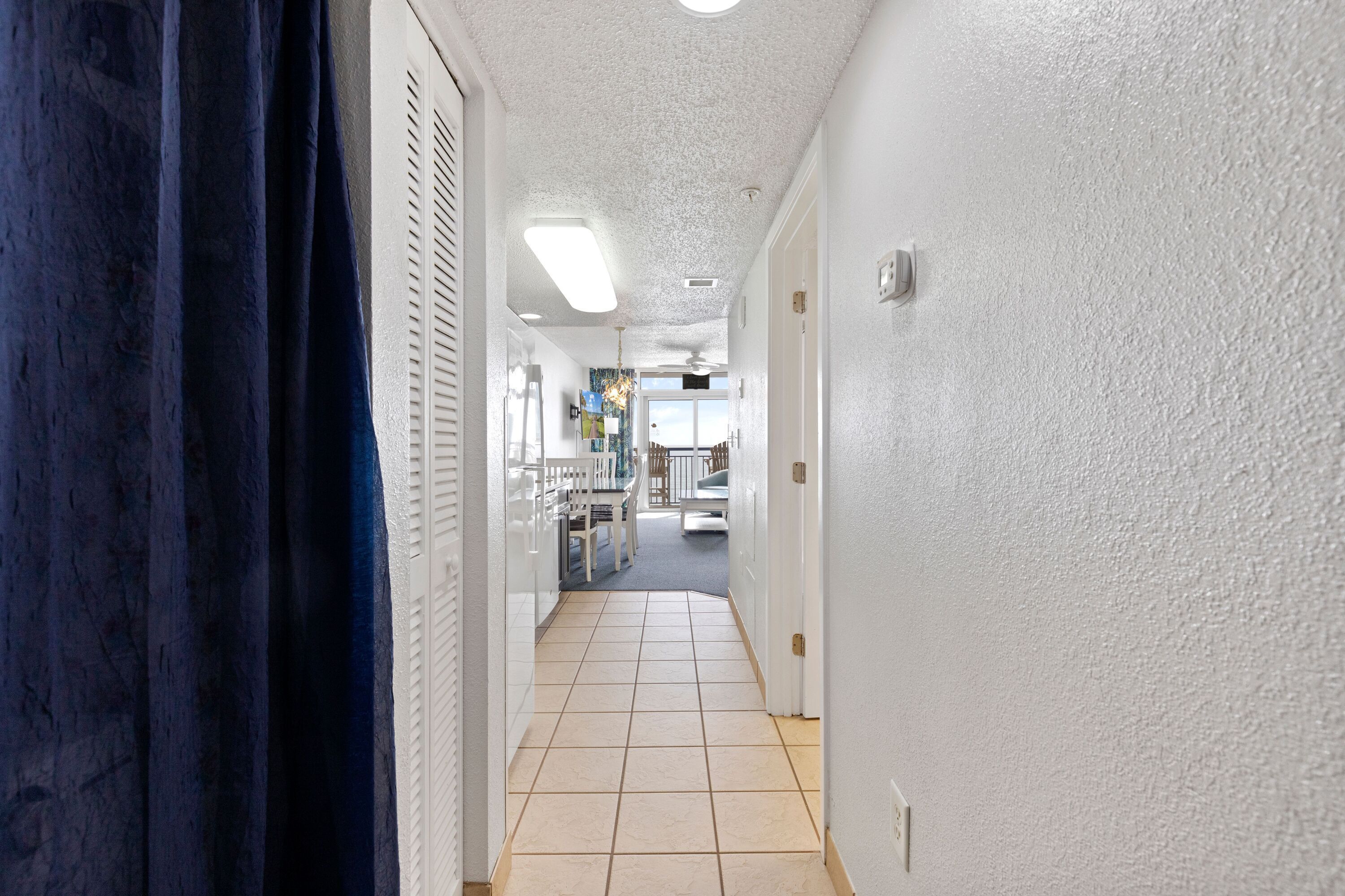 Un couloir aux murs blancs, doté d’un sol carrelé, et d’où l’on aperçoit une salle à manger avec une table et des chaises.