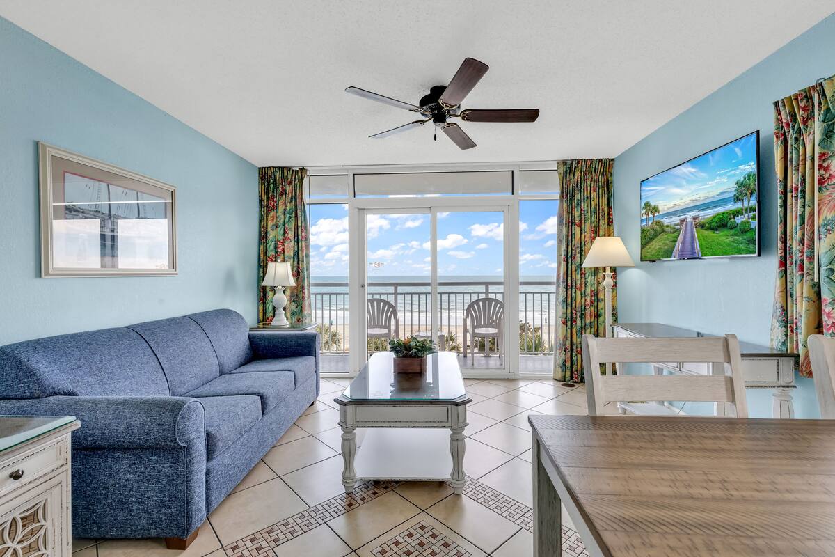 A living room with a blue sofa, a coffee table, a TV, and a balcony with a view of the ocean.