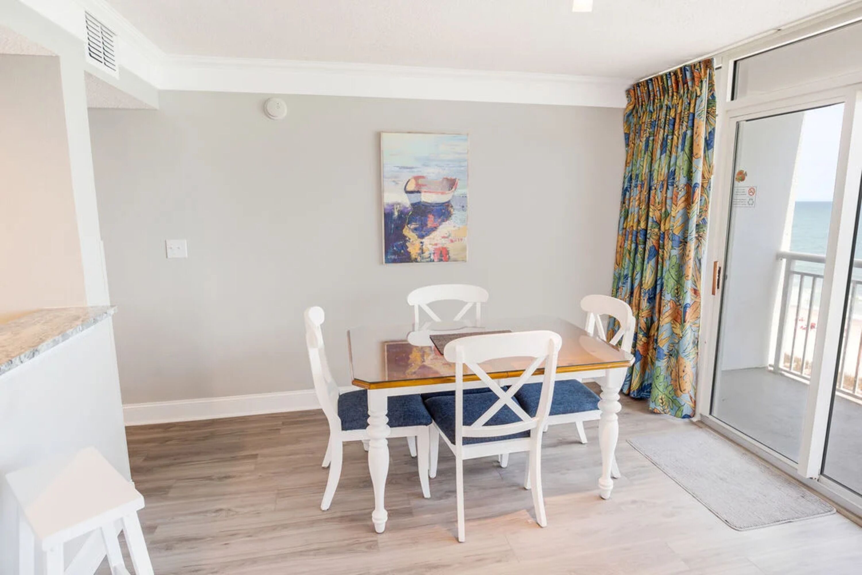 Un coin repas avec une table en bois et des chaises blanches, un tableau accroché au mur et un rideau coloré sur la porte-fenêtre coulissante.