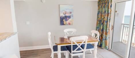 Un coin repas avec une table en bois et des chaises blanches, un tableau accroché au mur et un rideau coloré sur la porte-fenêtre coulissante.