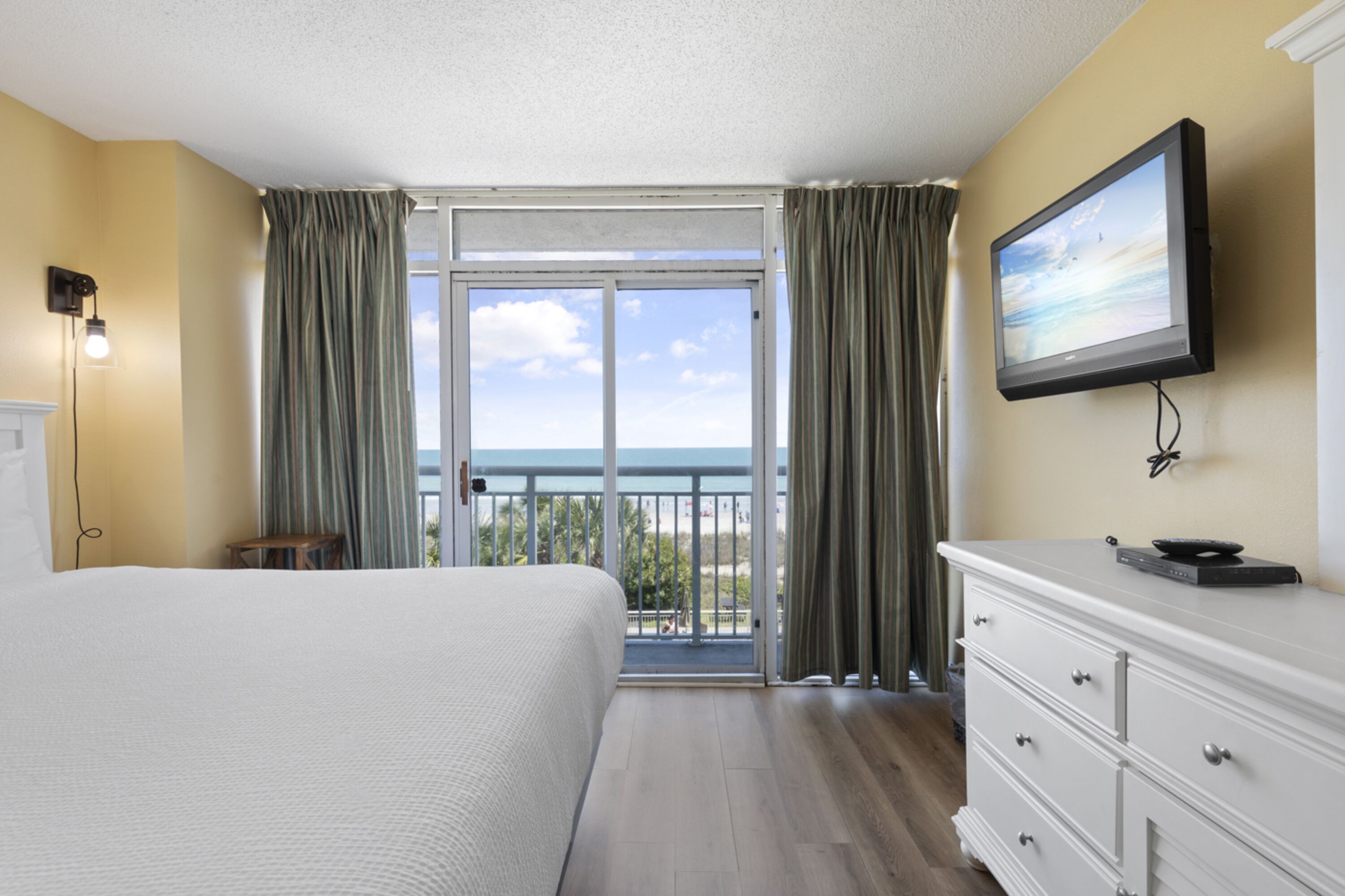 A hotel room with a large bed, a television on the wall, a dresser, and a balcony with a view of the sea.