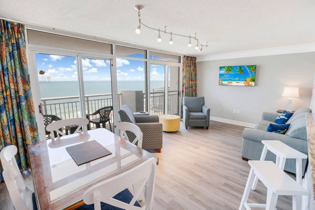 A spacious living area with a balcony overlooking the sea, a dining table, and a sofa.