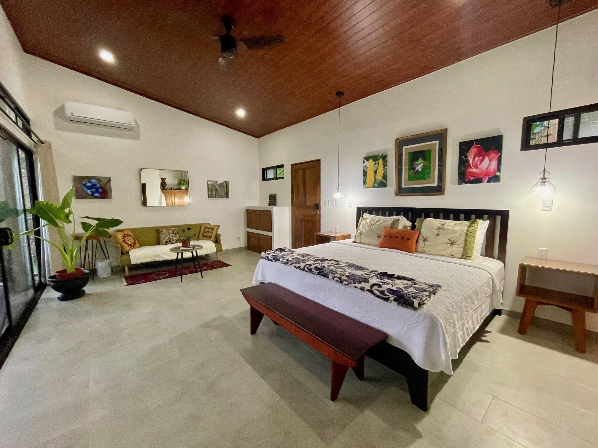 A bedroom with a large bed, a bench, a sofa, a small table, and a potted plant.