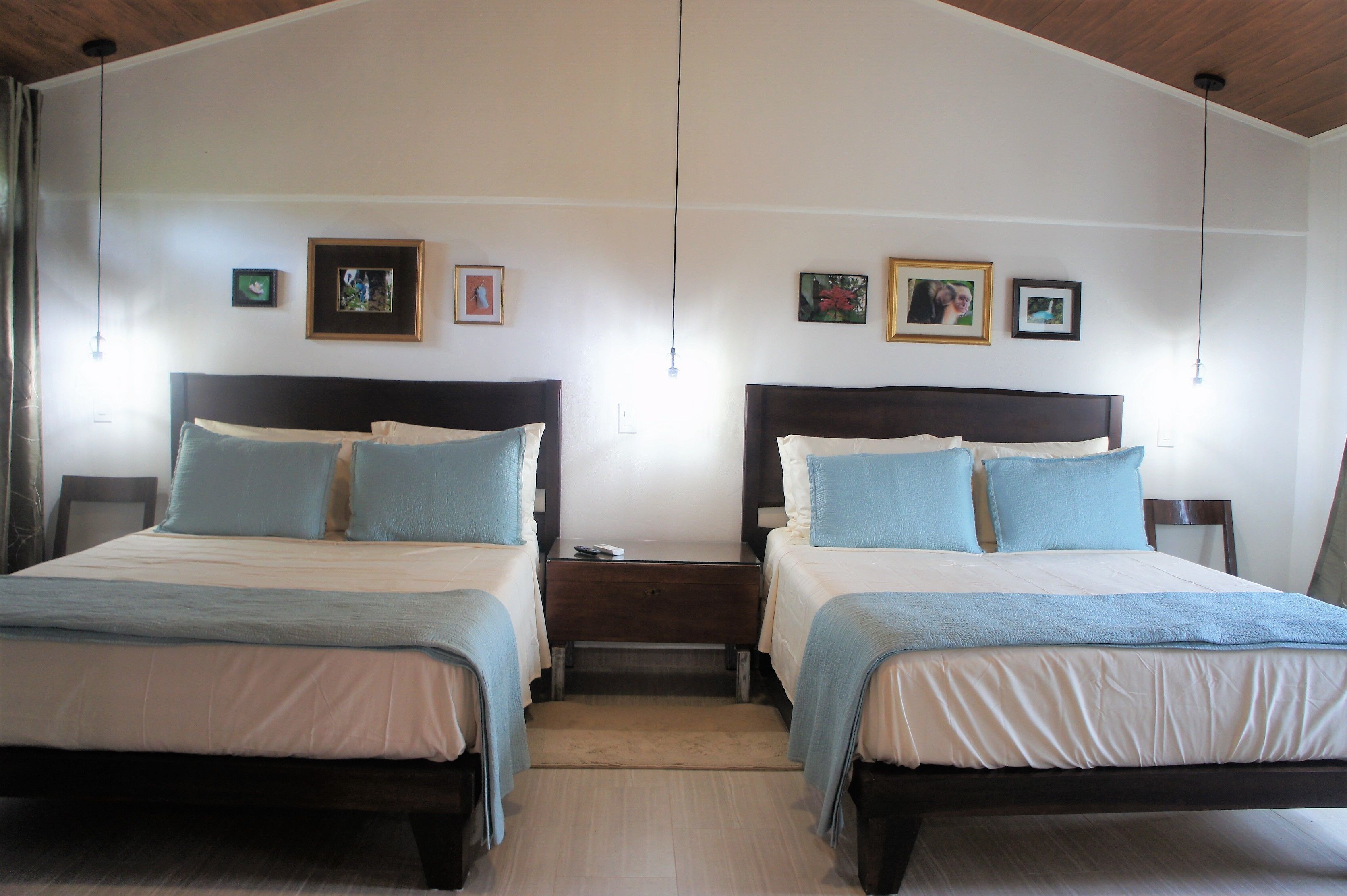 Habitación doble con dos camas, cada una con cabecero de madera, mesita de noche y una pequeña mesa. En la pared hay cuadros enmarcados.