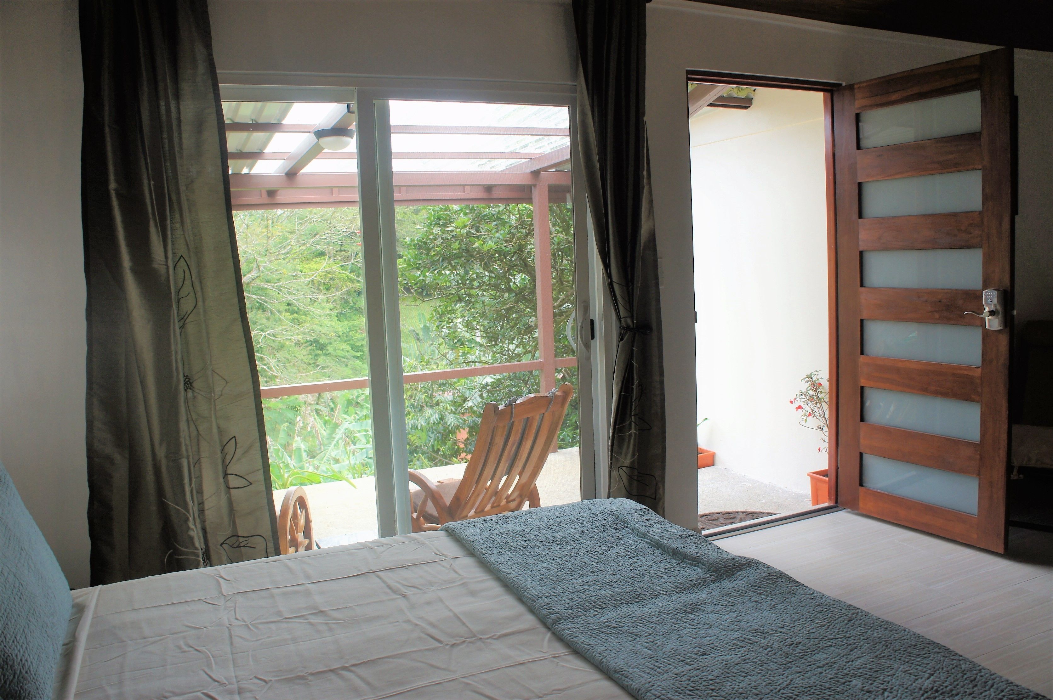 A bedroom with a bed, a wooden chair, a sliding glass door, and a view of greenery outside.