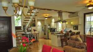 Uma cozinha espaçosa com uma ilha central, uma sala de jantar com mesa e cadeiras vermelhas, e uma sala de estar com sofá e apoio para os pés.